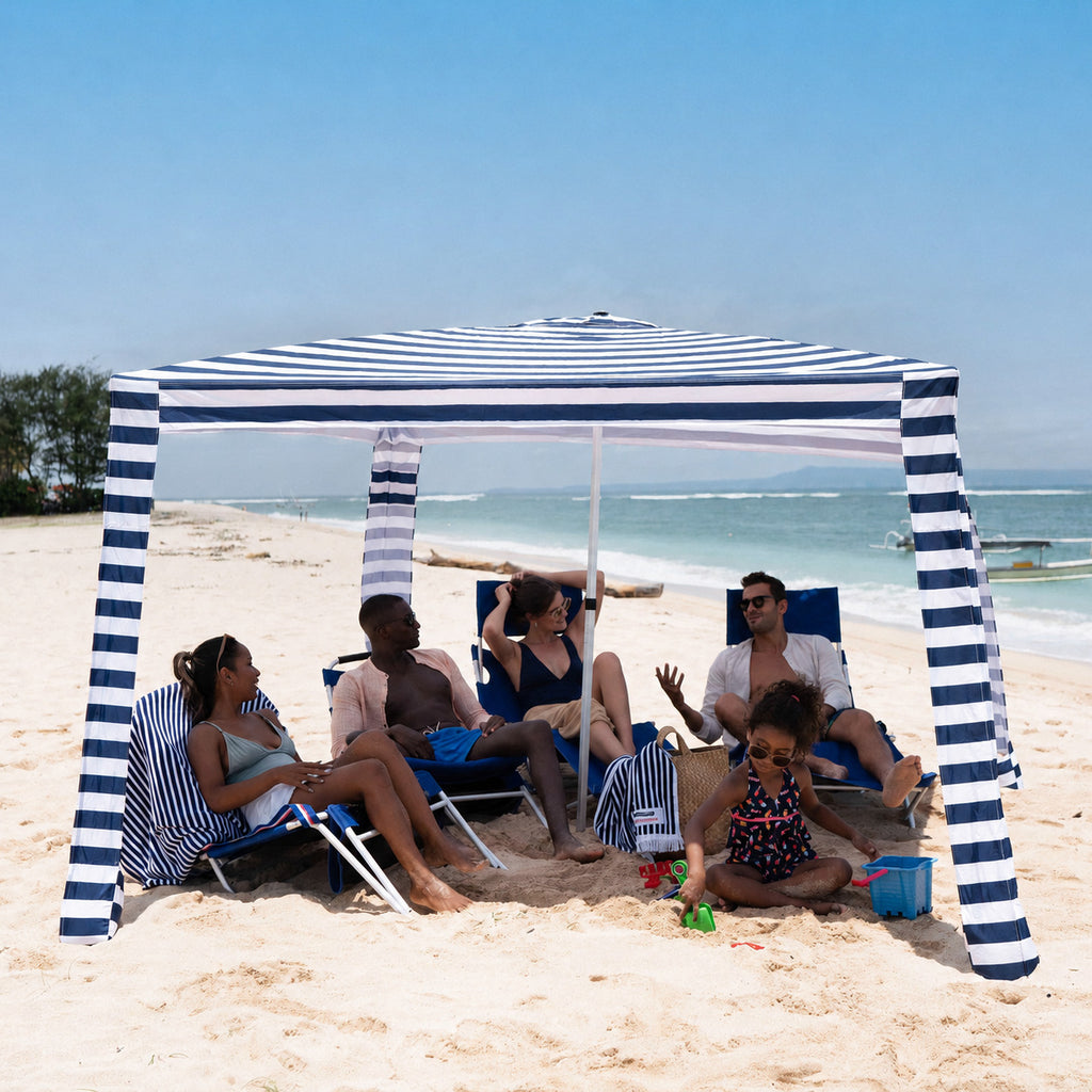 People sitting under a blue and white striped beach canopy on a sandy beach with ocean view.