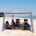People sitting under a blue and white striped beach canopy on a sandy beach with ocean view.