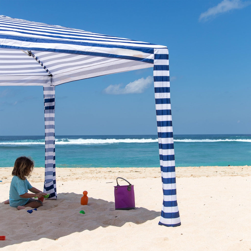 Child playing under a blue and white striped canopy on a sandy beach with ocean view.