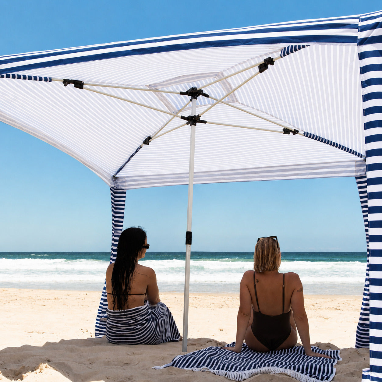 Two women sitting under a striped beach umbrella on a sandy beach with ocean view.