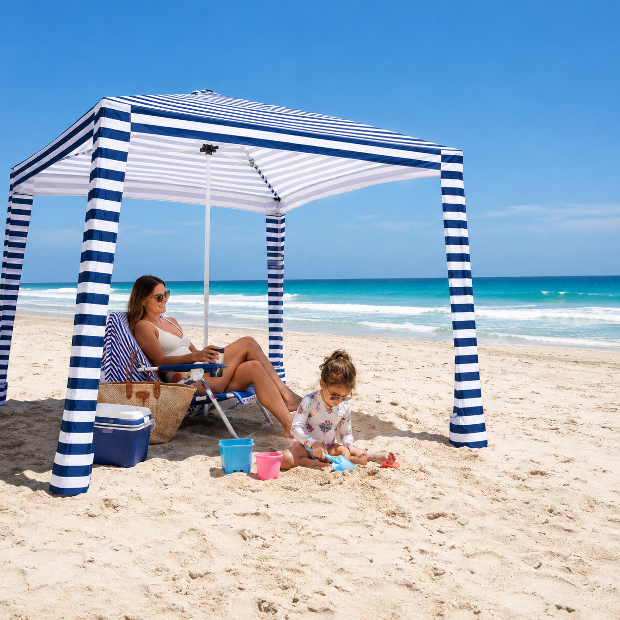Woman and child under a striped beach canopy on a sandy beach with ocean view.